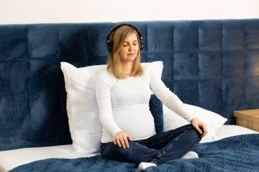 Pregnant woman meditating on bed with headphones.