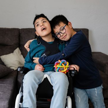 A boy in glasses affectionately hugs a boy in a wheelchair holding a colorful toy.