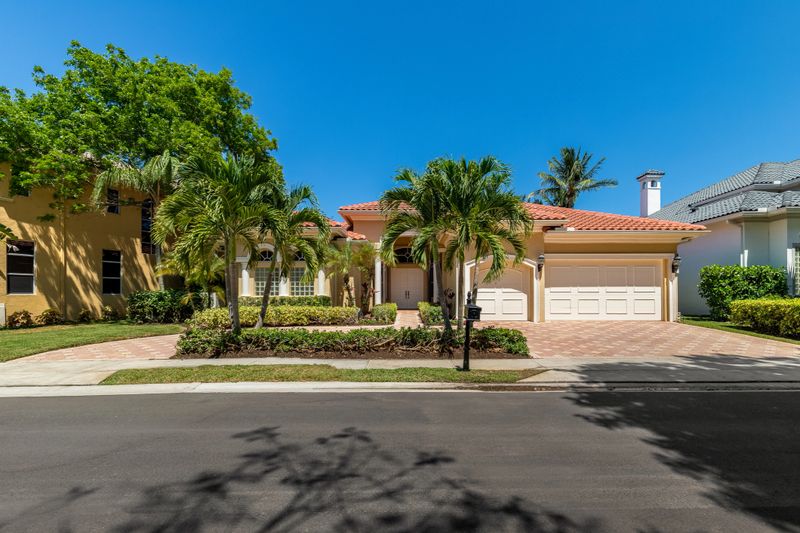 Facade of elegant colonial-style mansion in Boca Raton, with tropical front garden, cobblestone driveway, palms, trees, short grass, sidewalk, blue sky