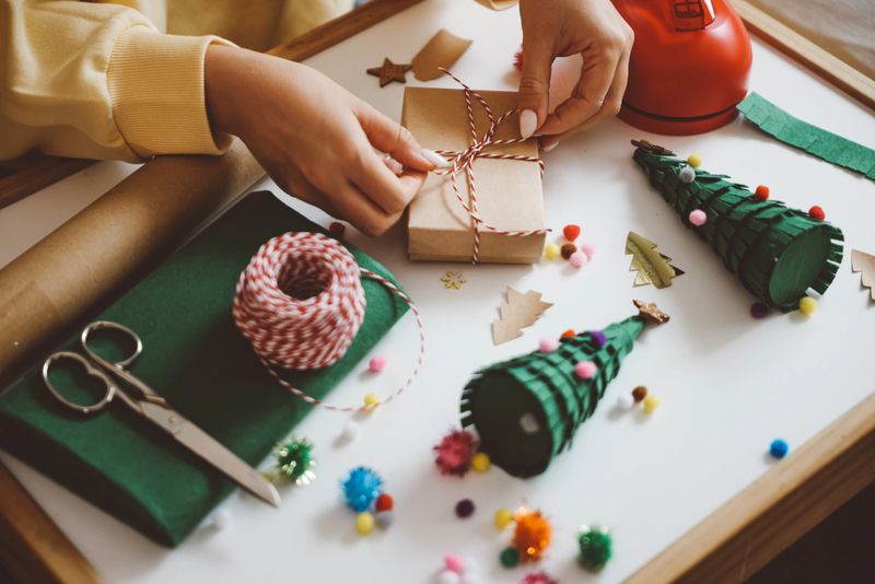 Young Woman Wrapping Christmas Gifts In Living Room. Creative diy craft hobby. Making handmade craft christmas ornaments and balls with felt spruce tree. Woman's leisure, holiday decorations. Closeup of female hands at white wood background