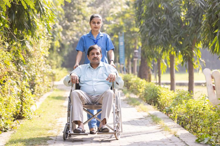 A caregiver pushes a man in a wheelchair along a sunlit garden path.