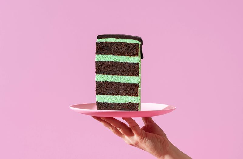 Woman's hand holding a plate with a slice of peppermint cake against a purple background. Homemade layer cake with mint-flavored cream and chocolate topping