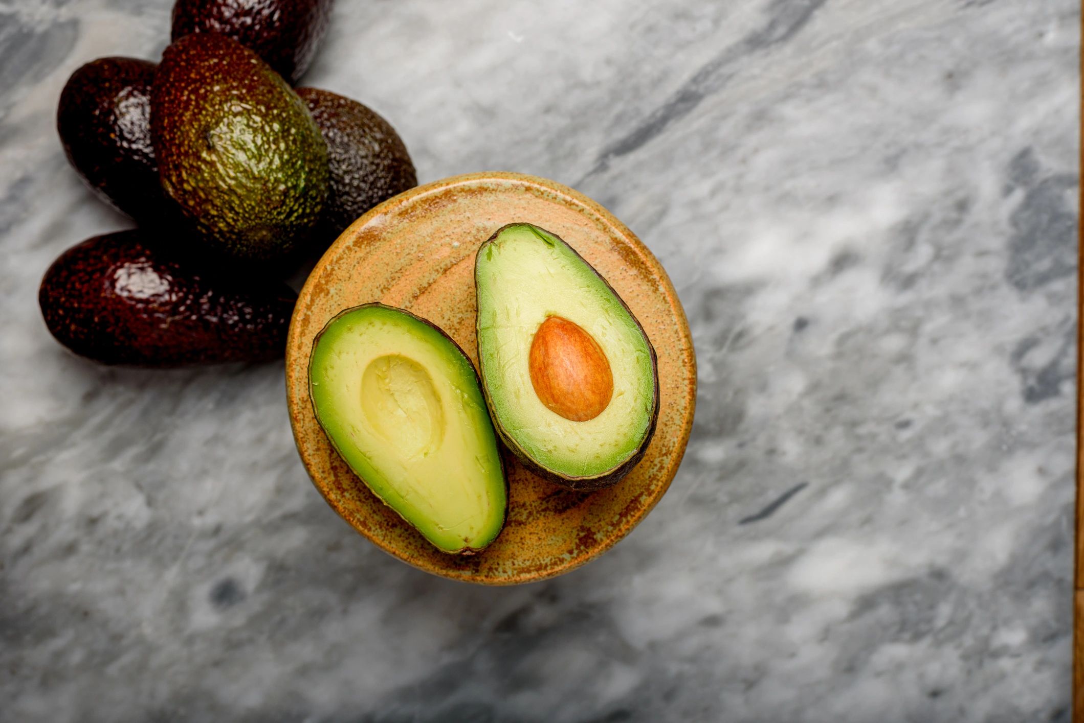 Halved avocado on a plate with whole avocados nearby.