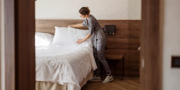 A maid in uniform makes a bed, adjusting white pillows