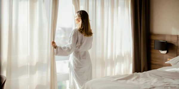 Woman in a robe looking out a window in a bedroom.