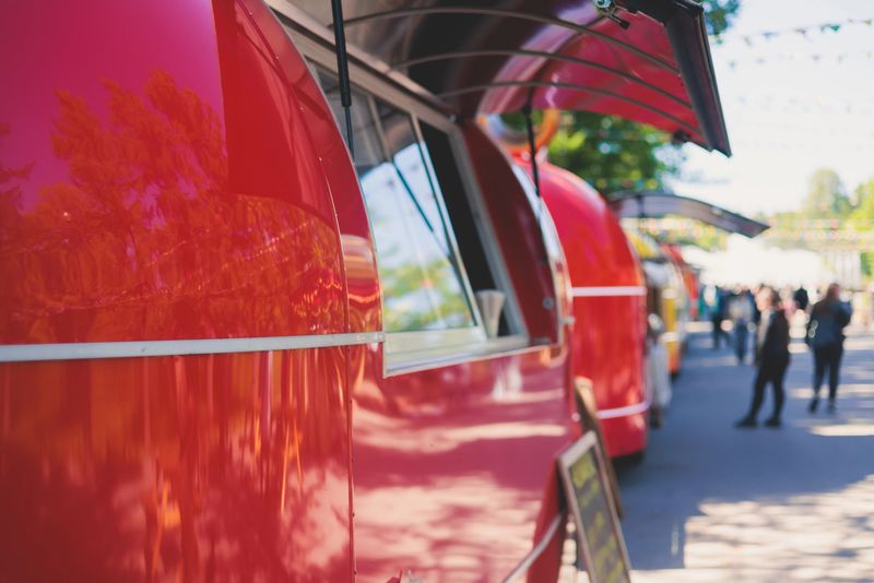 Girl choosing and ordering street food in colourful food truck van on a festival in a city park, summer sunny day, food stall kiosk offers fast food, coffee and drinks for sale, food court outdoors