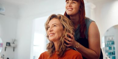 A hairstylist smiling while working on a client's hair in a bright salon.