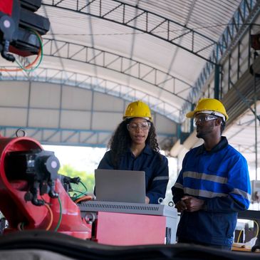 Two engineers in safety gear working on a laptop near industrial machinery.