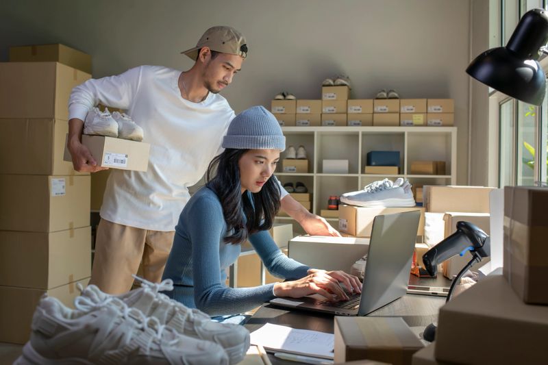 Asian woman and man online seller prepare parcel box and check orders of product for deliver to customer. Couple e-commerce business owner holding packages in store warehouse, online shopping concept.