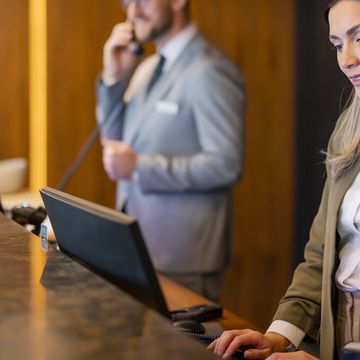 Two hotel receptionists assisting guests, one on the phone and one at the computer.