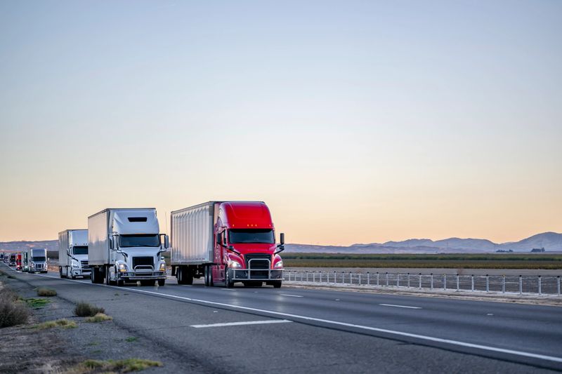Convoy of a industrial grade commercial professional transportation of different big rig semi trucks with semi trailers driving on the straight highway road at twilight time in California