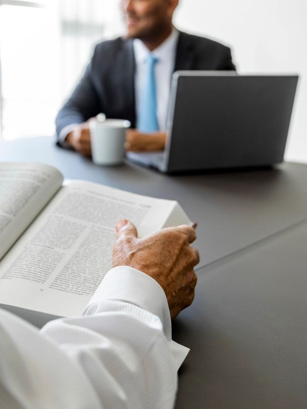 Older person reading a book while a younger man sits with a laptop and coffee.