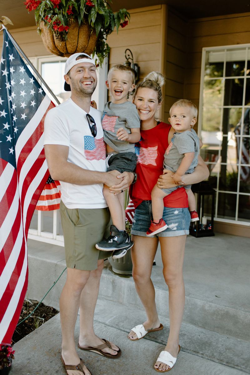 Portrait of a happy mom and dad holding their toddler boys and smiling directly at the camera while posing together on the front porch of their home on the Fourth of July.
