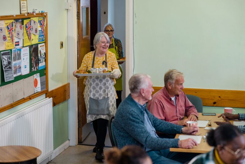 A wide shot of a medium group of people visiting a warm hub/food bank which is a safe place for people to enjoy a warm and friendly environment in the community with the current cost of living increasing. A volunteer wearing an apron is serving visitors roast dinners.