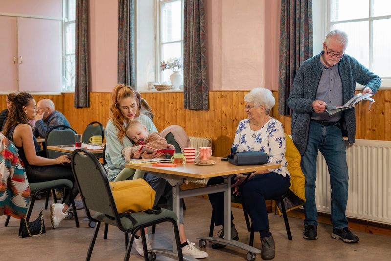 A shot of a small group of people visiting a warm hub/food bank which is a safe place for people to enjoy a warm and friendly environment in the community with the current cost of living increasing. A group of visitors are sitting at a table enjoying tea, biscuits and each other's company.