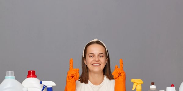 Smiling woman in orange gloves with cleaning supplies pointing up.