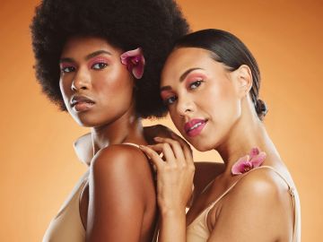 Two women with flowers in their hair pose against an orange background.