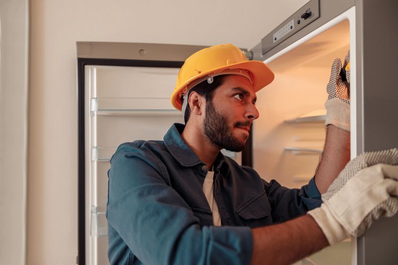 Male technician in uniform with screwdriver repairing refrigerator in kitchen. High quality photo