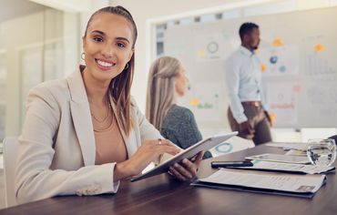 Smiling businesswoman using a tablet in a meeting room with colleagues in the background.