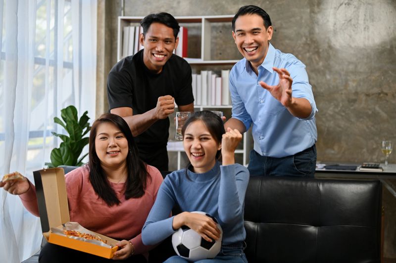 Group of joyful and excited Asian friends sit on sofa watching and cheering soccer or football team on TV together. sports and entertainment concept