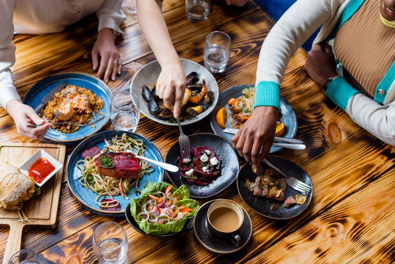 Unrecognisable friends sitting at a wooden table in a modern restaurant, enjoying a meal out together in Amble, North East England. They are all eating from the plates on the table.