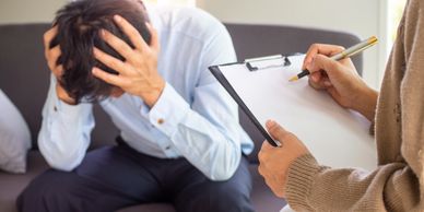 A distressed man holds his head while a therapist takes notes.