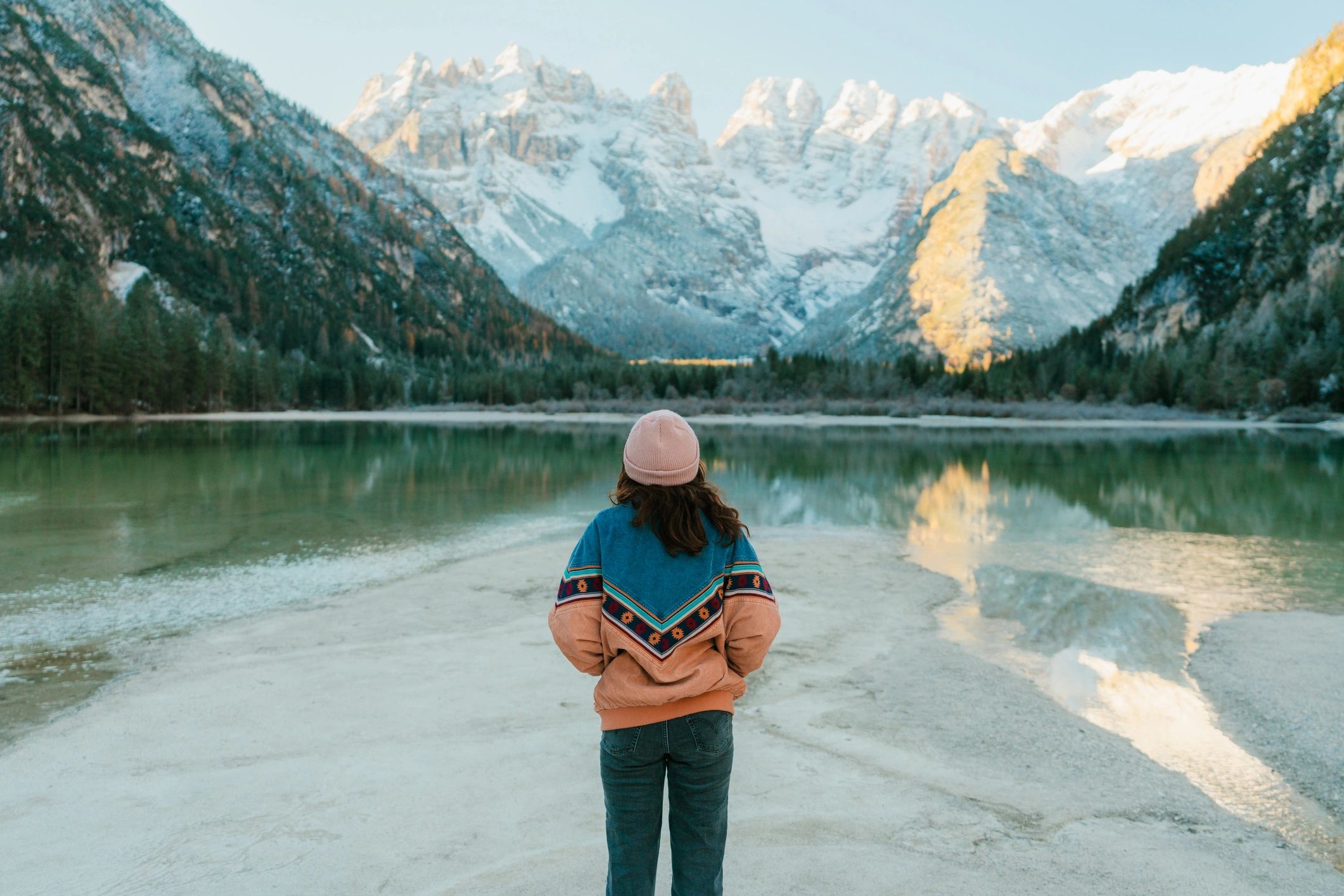 Person standing by a serene mountain lake with snow-capped peaks.