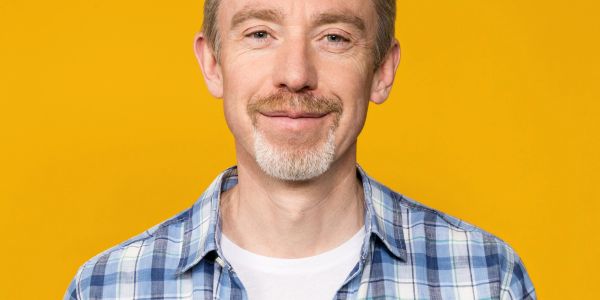 Smiling man in a blue plaid shirt against a yellow background.