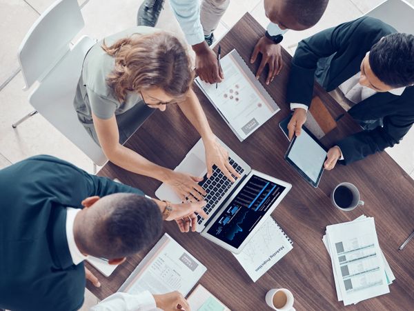 A team collaborates around a table with laptops, tablets, and reports.