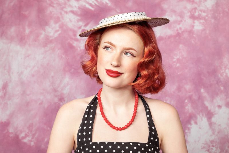 Close up studio portrait of a cheerful red-haired white woman dressed in retro style in a polka-dot dress against a pink background
