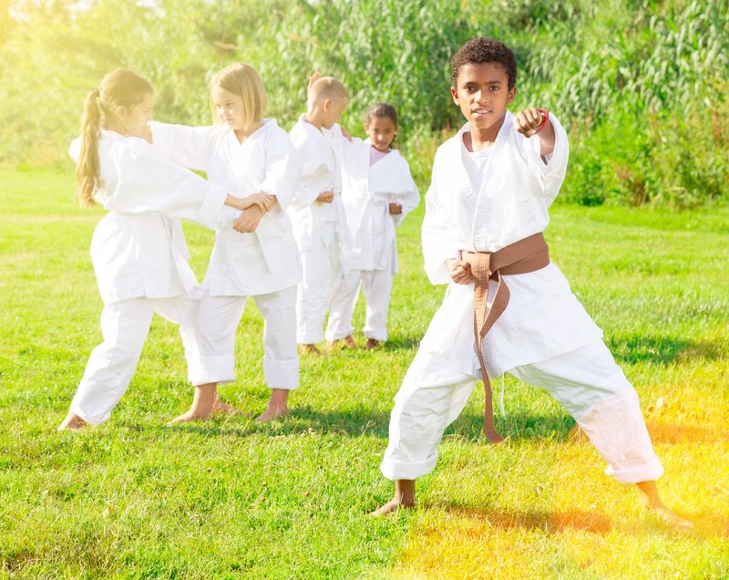 Positive afro american schoolboy in kimono with white belt practicing karate with his friends in park on sunny day