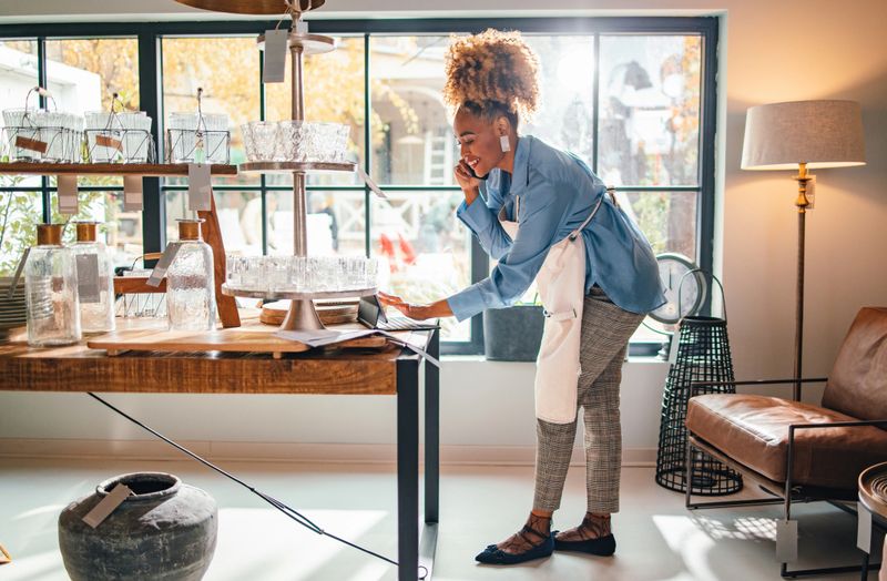 Happy African-American businesswoman in apron typing online order on a digital tablet while speaking on the phone with a client and standing in the store.