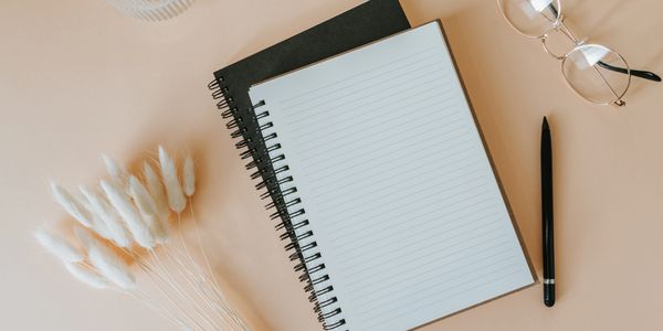 Open notebook, pen, glasses, and dried flowers on beige surface.