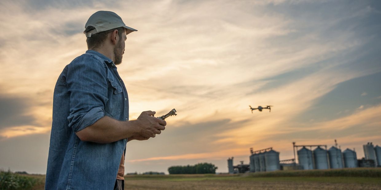 A man operates a drone over a field at sunset near industrial silos.