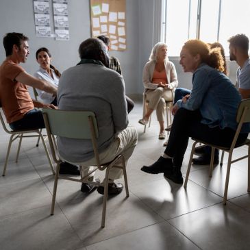A group of people engaged in a serious group therapy session in a bright room.