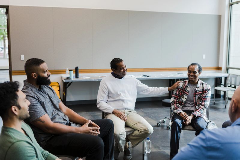 The man places his hand on his friend's shoulder while he tells a positive story about his home life improving since coming to meetings.