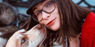 A woman with glasses and a hat is affectionately kissed by a dog.