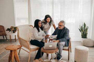 Three people discussing documents in a cozy living room with a laptop.