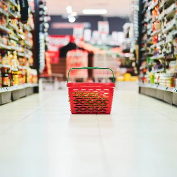 A red shopping basket sits empty in a supermarket aisle.