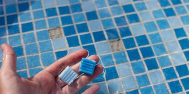 Hand holding two blue dishwasher detergent pods over a tiled surface.