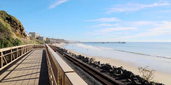 Scenic coastal boardwalk with railroad tracks and a distant pier under a clear blue sky.