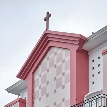 Top section of a pink church facade with a cross against a cloudy sky.