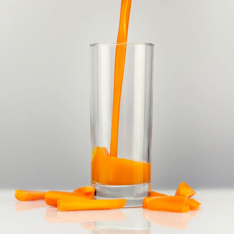 Fresh carrot juice is poured into a glass, which stands on the white table with pieces carrot. Healthy vegan food. A natural product containing vitamin A. Selective focus, front view