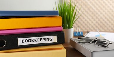 Stack of colorful binders with a 'BOOKKEEPING' labeled binder on a wooden desk.