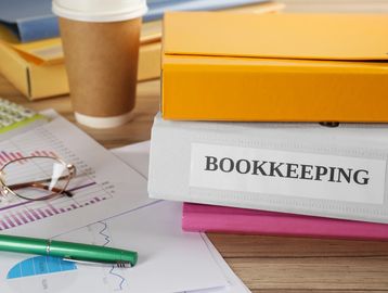 Stacked binders, papers, and a coffee cup on a desk for bookkeeping.