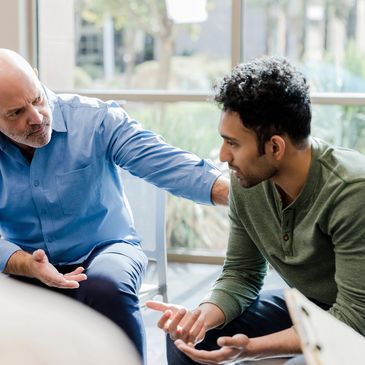 An older man comforts a younger man during a serious conversation in a bright room.