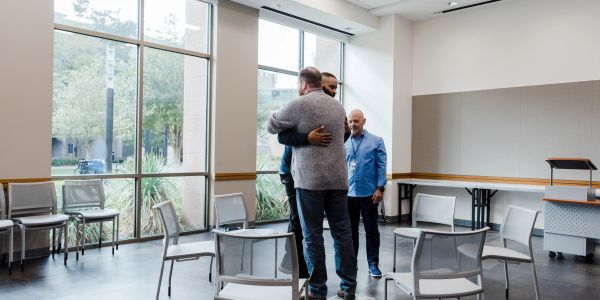 Two men hugging in a bright room with chairs.