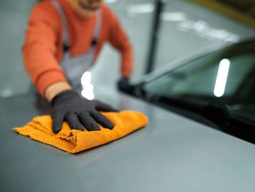 Person cleaning a car hood with an orange cloth wearing black gloves.