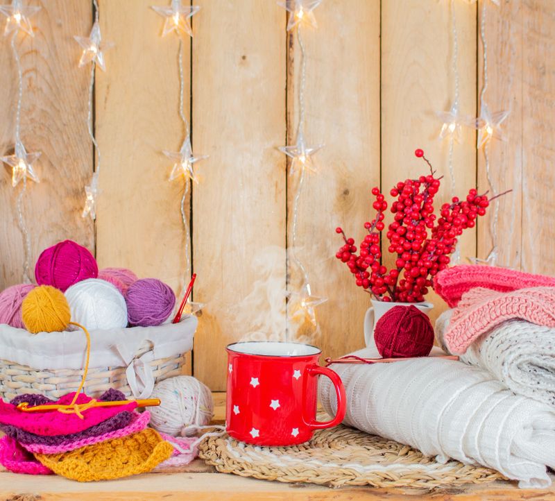 a red mug with hot drink, colorful woolen balls in a basket and granny squares, crochet hooks, lights and woolen blanket in front of a light brown wooden wall