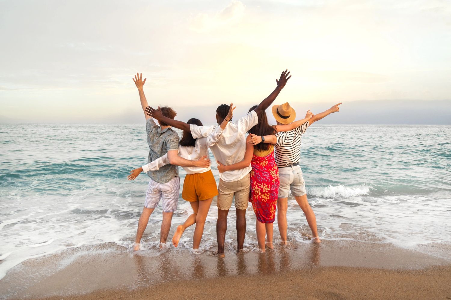 A group of friends joyfully standing on the beach with arms raised at sunset.